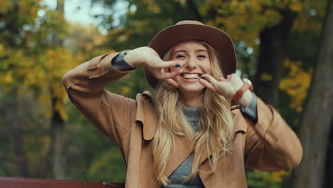 caucasian young blonde woman in a hat and coat laughing while posing to the camera and doing funny muzzles and gestures in the park in autumn