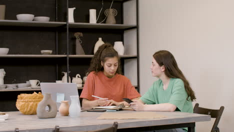 pottery studio owner talking with a client and taking notes on a clipboard while client showing products on mobile phone