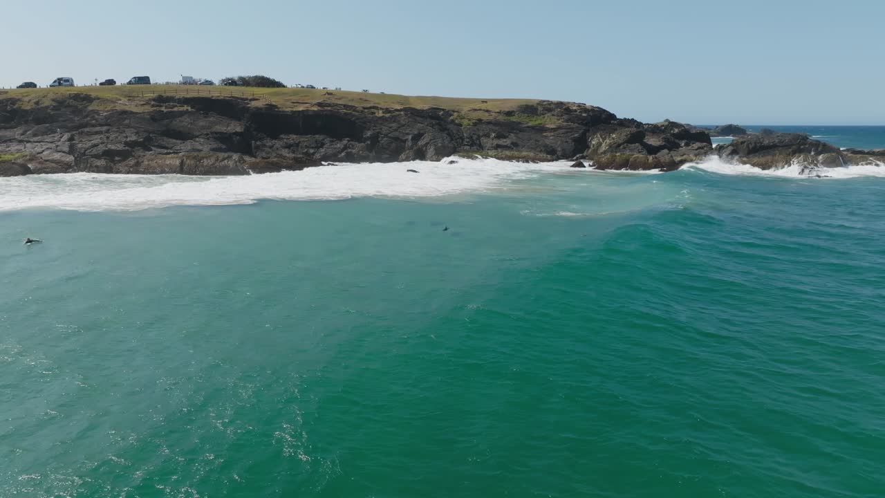 Drone clip of gentle swell rolling past rocky headland, with surfer and pod of dolphins in the bay
