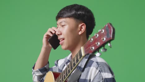 close up side view of asian teenager with guitar talking on smartphone while standing on green screen background