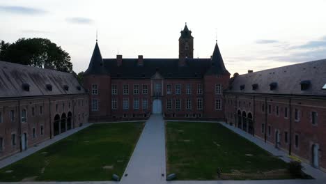 Aerial-circular-view-of-entrance-and-french-garden-of-Alden-Biesen-Castle-in-Belgium,-Germany-spread-across-large-area-with-greenery-and-landscape-at-sunset