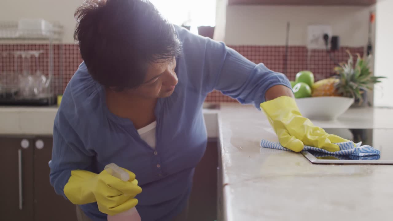 Mulher idosa birracial sorrindo, usando luvas e limpando a mesa na cozinha sozinha