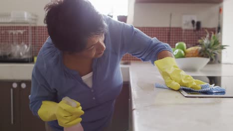 smiling senior biracial woman wearing gloves and cleaning table in kitchen alone