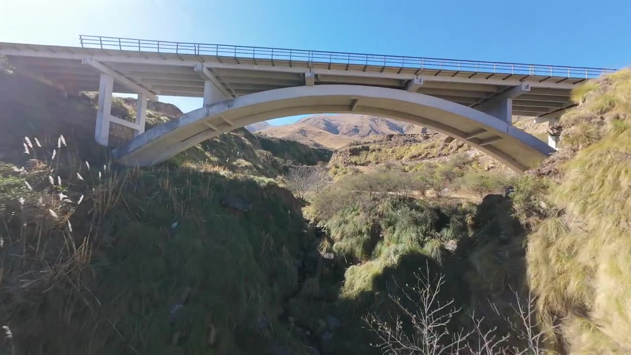 Dynamic FPV drone video diving into a canyon and river valley of Tafí del Valle, Tucumán, Argentina, gliding beneath a bridge and showcasing rugged terrain in crisp daylight.