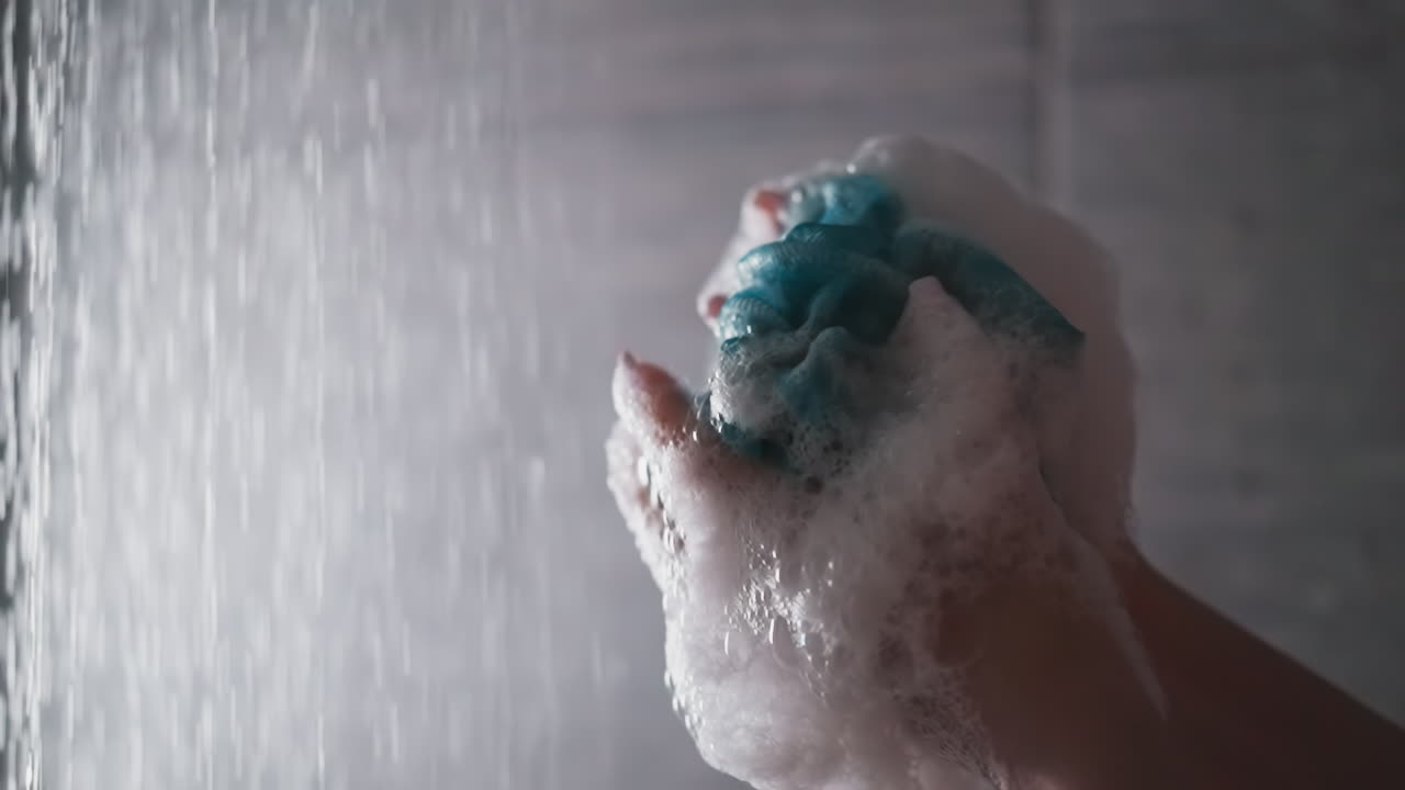 Woman crumples foamy loofah in shower cabin closeup. Lady shows mesh body washing sponge against falling water flow. Skincare and massage tool