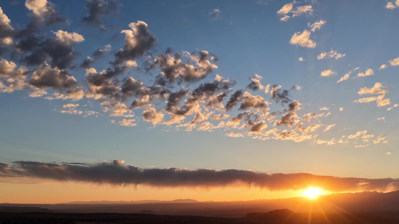 4K Aerial drone timelapse of the sun rising between a distant mountain range and a dark band of clouds. Golden morning rays brilliantly illuminate the textured, scattered clouds above.