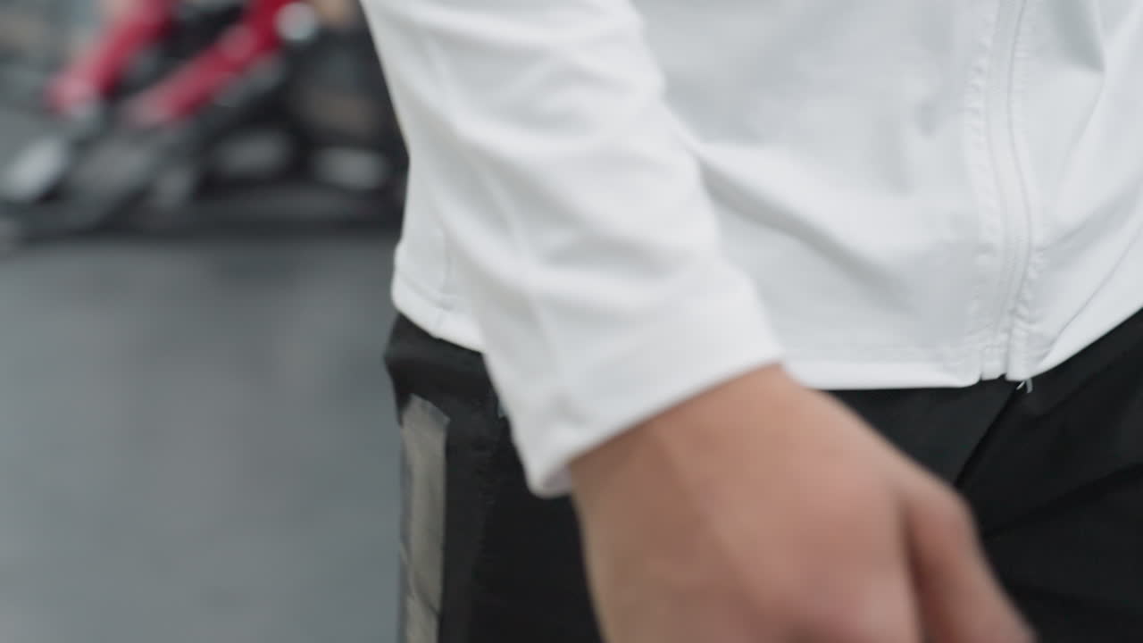 close up of man wearing black smartwatch and white athletic top carrying large leather duffel bag and holding wireless headphones while walking through bright modern gym corridor with equipment