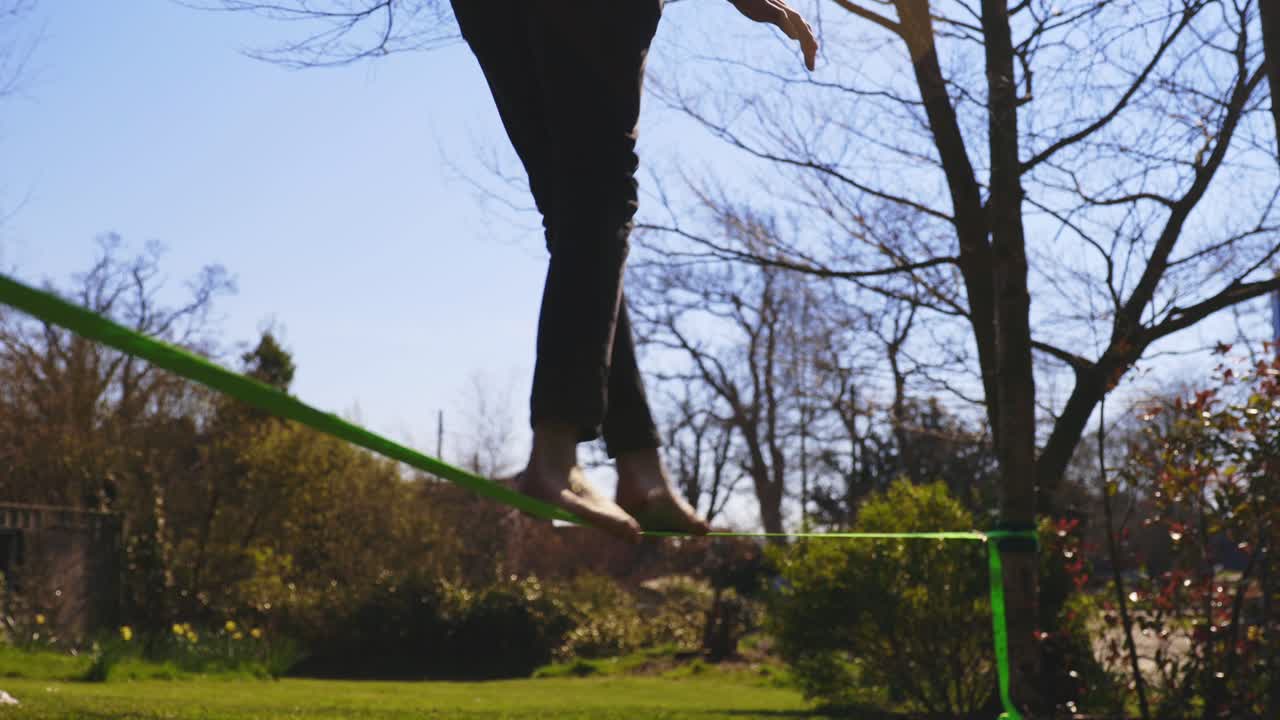 검은 청바지와 복어 코트를 입은 젊은 맨발의 낮은 각도에서 걷고 있다가 슬랙 라인에서 떨어짐