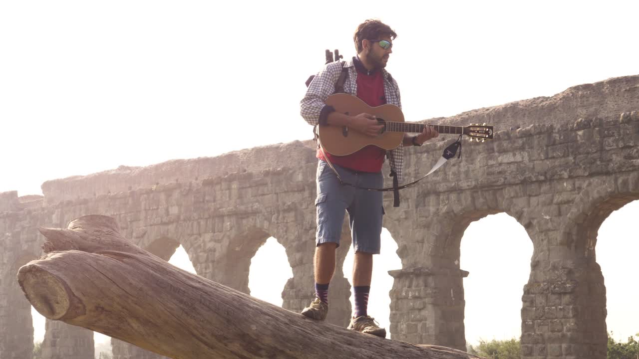joven aventurero viajero de pie en la parte superior de un tronco de tronco tocando la guitarra cantando frente a las antiguas ruinas del acueducto romano en el parque degli aquedotti en roma al amanecer en cámara lenta