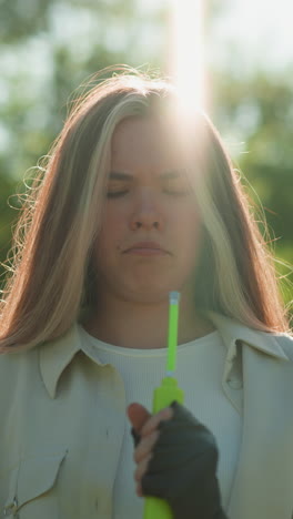 chica de pelo rubio de pie pensativa, sosteniendo una bomba de aire verde con expresión enfocada como la luz del sol crea un efecto radiante en su cabeza, en el fondo de árboles exuberantes