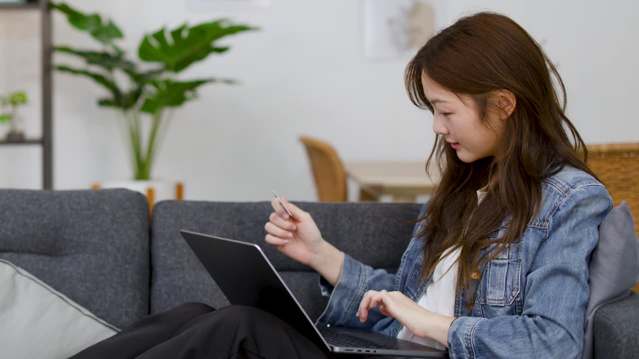 A young Asian woman sits on a sofa using a laptop and credit card indoors