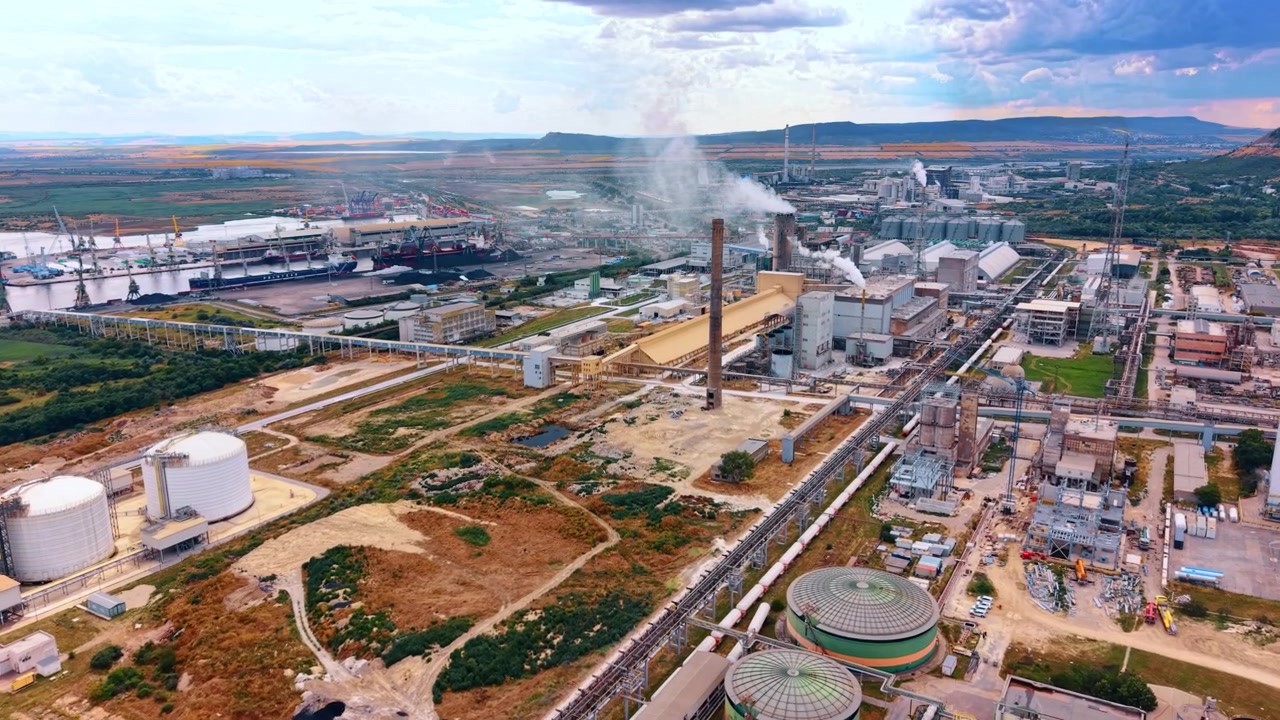 Industrial chemical refinery plant near a river terminal. White storage tanks and production units emit smoke within a large infrastructure zone next to distant water.