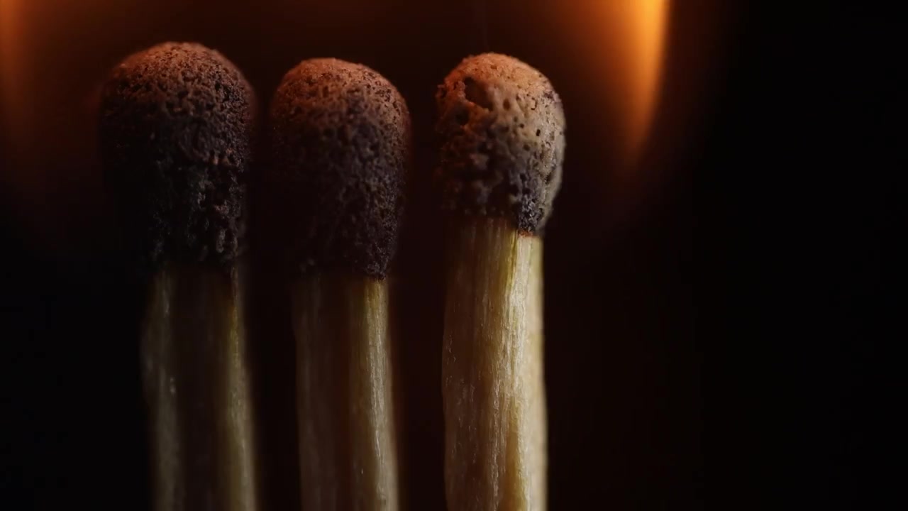 Un fiammifero di legno acceso brucia con una fiamma giallo-arancio brillante su uno sfondo nero profondo, creando un forte contrasto e un'illuminazione drammatica