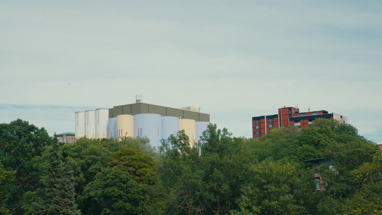 Grandi serbatoi industriali ed edifici di servizio si ergono sopra una fitta linea di alberi a Oslo, Norvegia, creando una scena urbana a strati dove infrastrutture, abitazioni e verde urbano si incontrano sotto un cielo tenue.