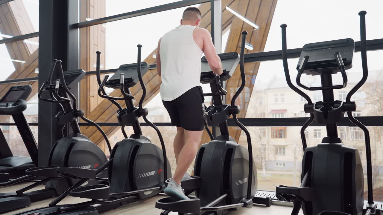 Trainer performs intense energetic elliptical workout in modern gym by tilted window showing urban apartments outside, stepping on pedals and adjusting console for dynamic cardio session