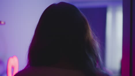back view of artist in fluffy sweater walking through doorway into vibrant neon lit studio featuring ring light, equipment and decor under pink purple ambient glow