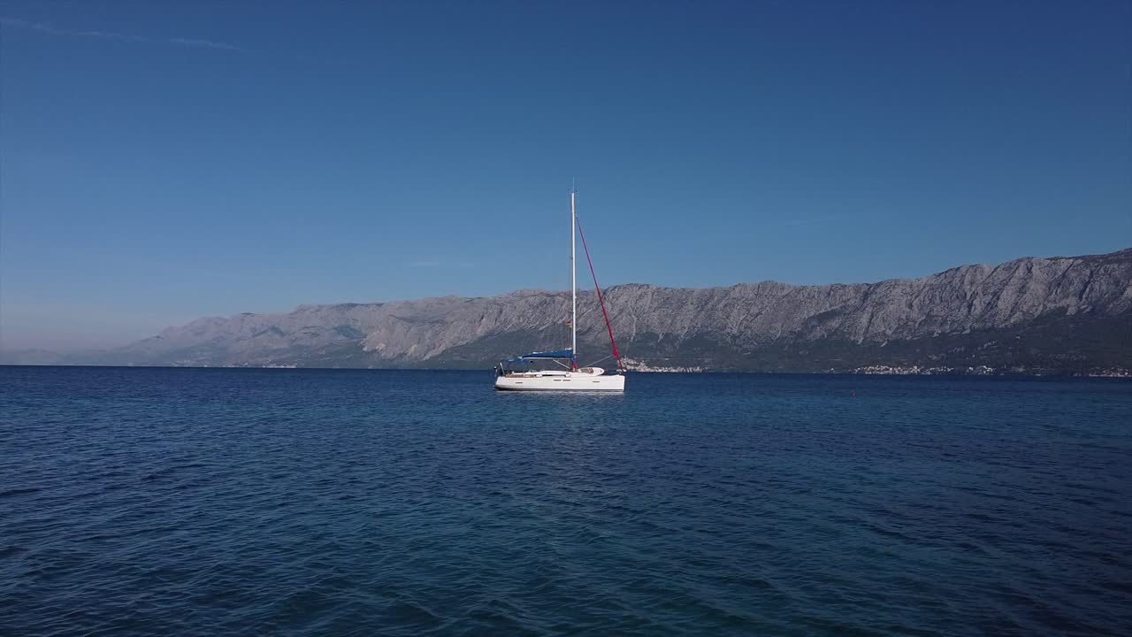 Drone che vola a bassa quota verso una barca a vela in una baia aperta in Croazia con le montagne sullo sfondo, sorvolando l'albero