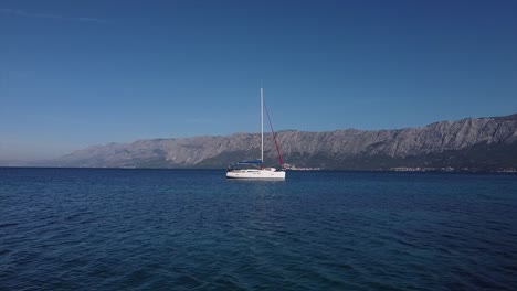 Drohne-Fliegt-In-Geringer-Höhe-Auf-Ein-Segelboot-In-Einer-Offenen-Bucht-In-Kroatien-Zu,-Mit-Bergen-Im-Rücken,-Die-über-Den-Mast-Fliegen