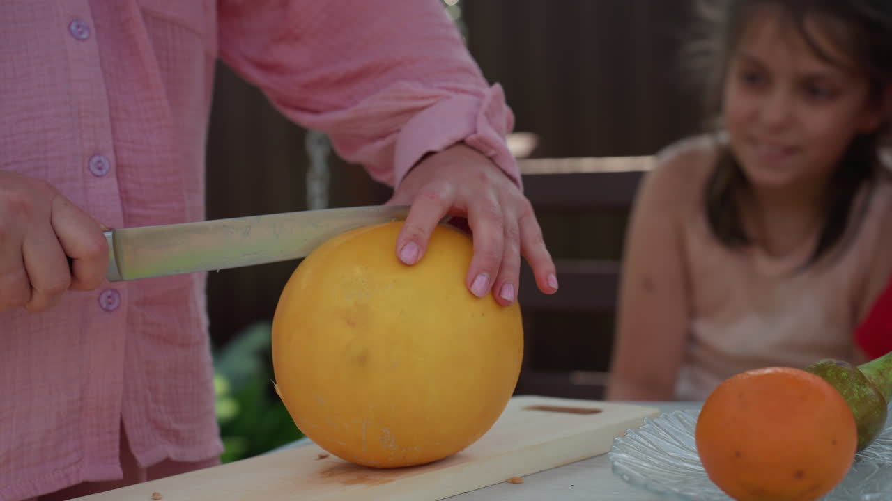 Caucasian Woman Slicing Melon Outdoors Girl On Wooden Table With Knife Cutting Ripe Flesh, Curious Daughter Watching, Orange On Plate, Summer Light, Familial Teaching Moment, Gentle Hands Guiding Safe