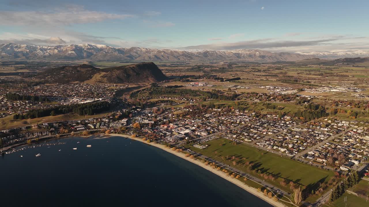 Wide panoramic aerial drone view of Wanaka town on lakefront with snow-capped Southern Alps mountain range in background, New Zealand