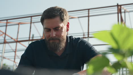 Close up bearded man working among strawberry plants under greenhouse frame, green leaf foreground in soft focus, sunlight, concentration, garden maintenance