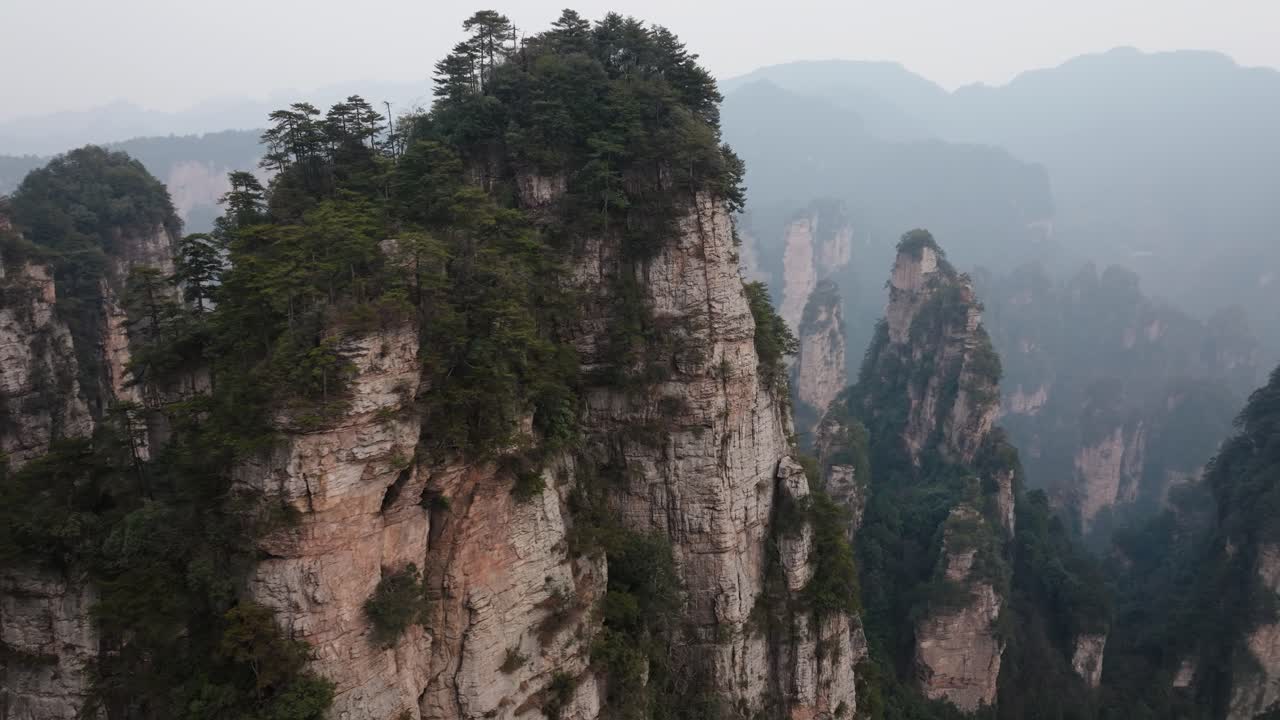 Orbiting aerial of a towering sandstone peak blanketed in trees with distant misty cliffs in Zhangjiajie National Park
