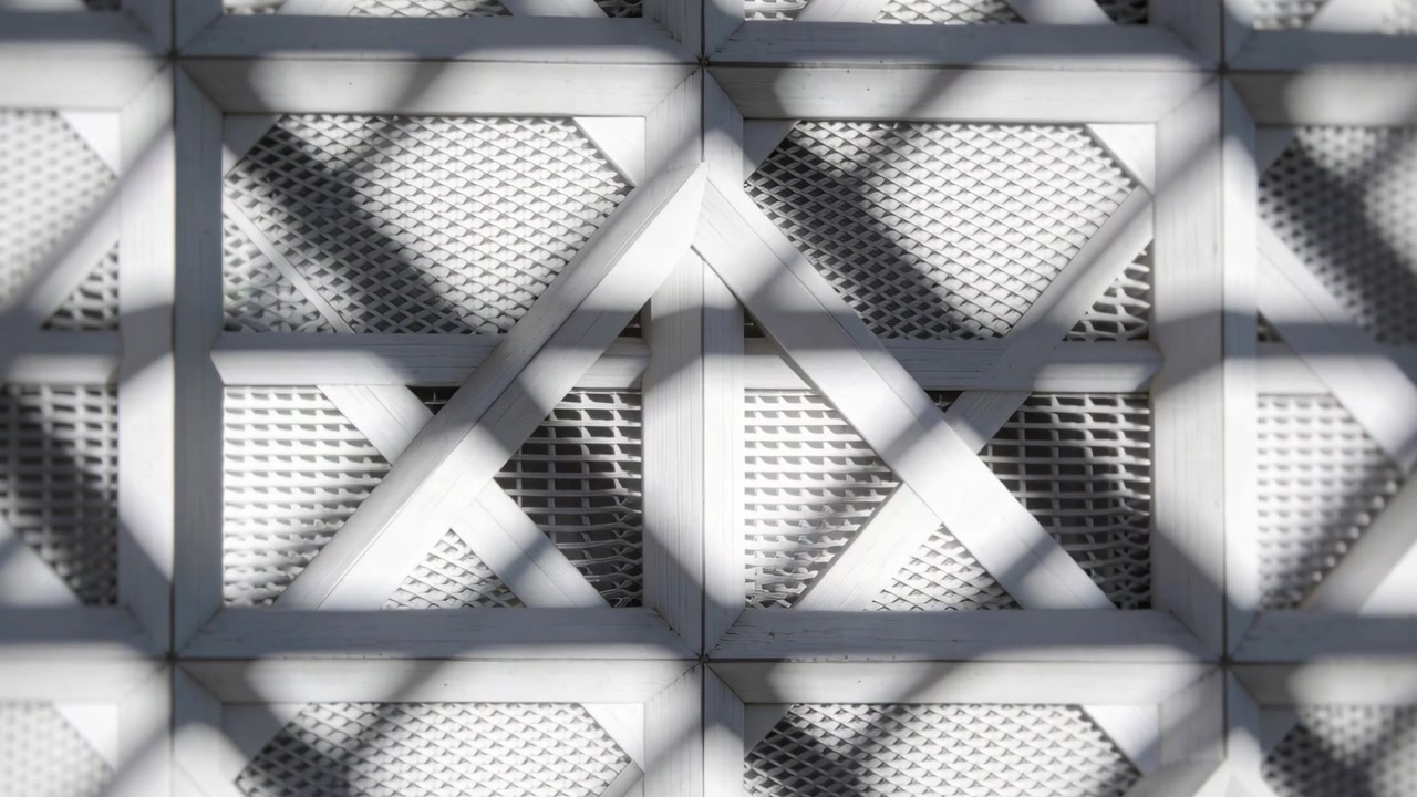 Luce solare mutevole che proietta ombre a reticolo attraverso pannelli modulari bianchi della facciata, rivelando una rete metallica.