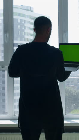 man looking out window at city view with laptop