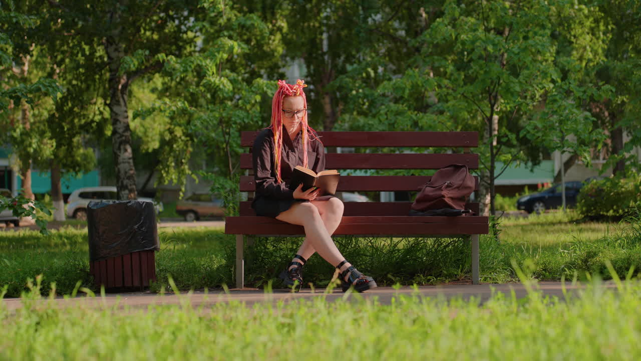 분홍색 땋은 머리를 한 여성이 야외에서 학습 자료에 집중하고 있어요. 활기찬 분홍색 땋은 머리를 한 젊은 백인 여성이 은은한 도시 공원에서 여름 동안 독서와 필기를 하고 있어요.