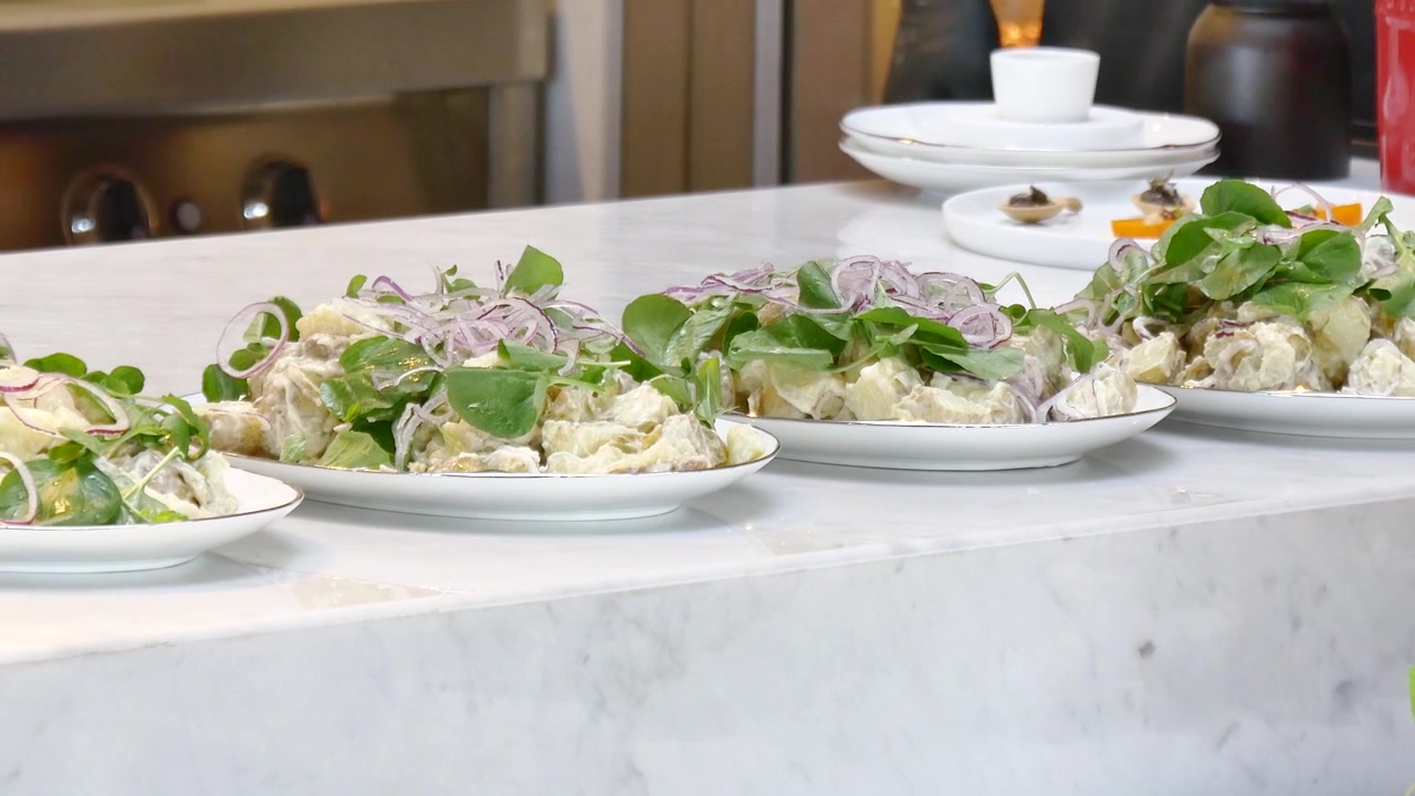 Insalate di patate cremose appena preparate, guarnite con crescione croccante e cipolle rosse affettate sottilmente, elegantemente impiattate su piatti di ceramica bianca lungo un bancone di marmo per catering professionale