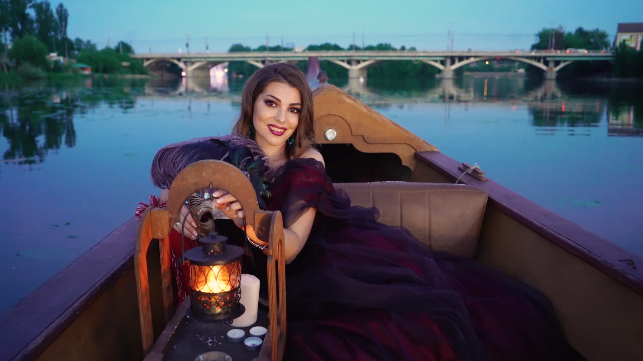 An elegant woman in a burgundy long dress is touching a decorative lantern and smiling on the gondola on the background of the river in the evening. Carnival