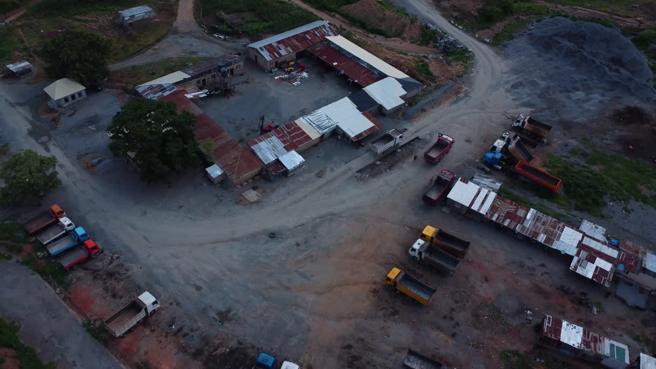 Dusk aerial footage of a bustling industrial quarry site with trucks, buildings, and machinery. Drove captures a moving granite truck driving through a rugged terrain on a narrow path in Abuja Nigeria