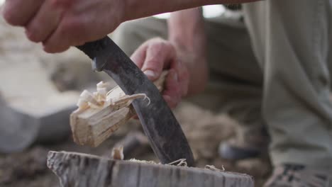 caucasian male survivalist using machete to prepare tinder and kindling wood at camp in wilderness