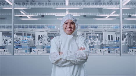 científico masculino asiático cruzando los brazos y sonriendo en una fábrica de laboratorio moderna con robots automatizados