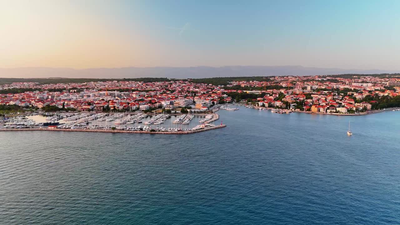 Aerial drone footage following the coastline in Zadar, Croatia—gliding over a large marina and warm red rooftops of the city as golden evening light sets over the Adriatic Sea.
