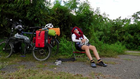 a young asian woman resting close to roadside trees shadow on a foldable light weight travelling chair beside her foldable bicycle loaded with waterproof backpacks after a hectic ride, thailand