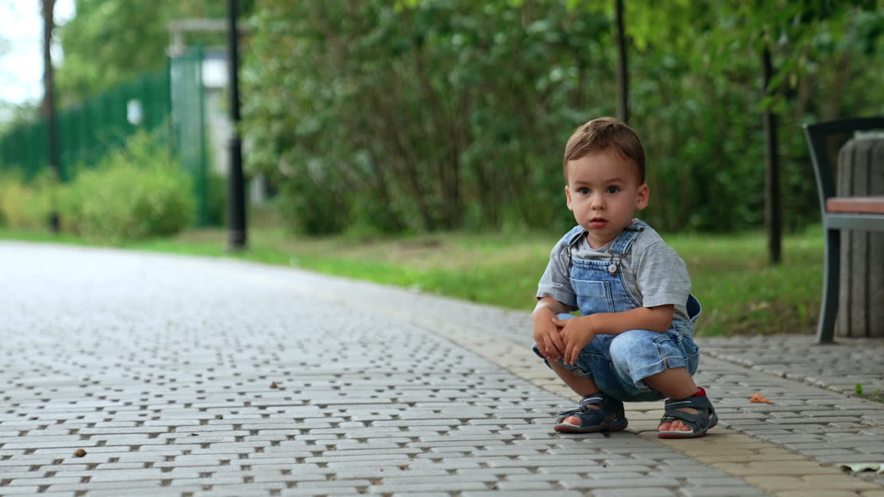 Beautiful Caucasian kid sits squatted on the paved road. Kid looks thoughtfully ahead and then runs.