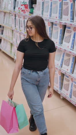 vista trasera de una mujer joven con camiseta negra y vaqueros caminando por el pasillo de la tienda, navegando por productos de cama mientras sostiene bolsas de compras, otro comprador pasa en el fondo