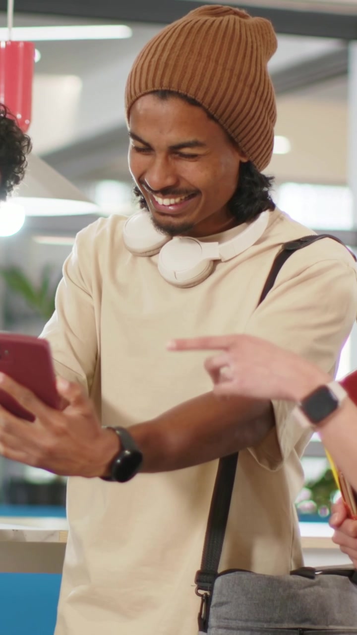 Vertical video: Reaching across to left friend, man with beanie holding phone in lounge for selfie
