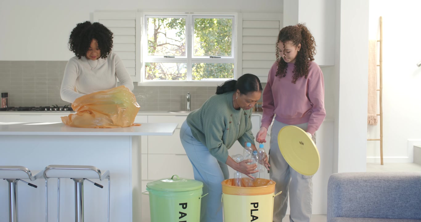 African American female family sorting recycling in kitchen with bins after senior holding bottles
