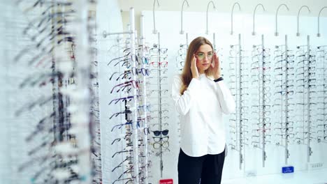 optical store. woman choosing spectacles indoors. young girl in white blouse trying on new eyeglasses on stylish glasses background. eyesight concept.