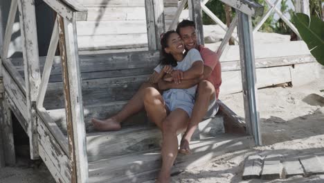 Happy-diverse-couple-embracing-sitting-on-the-steps-of-wooden-beach-house,-in-slow-motion