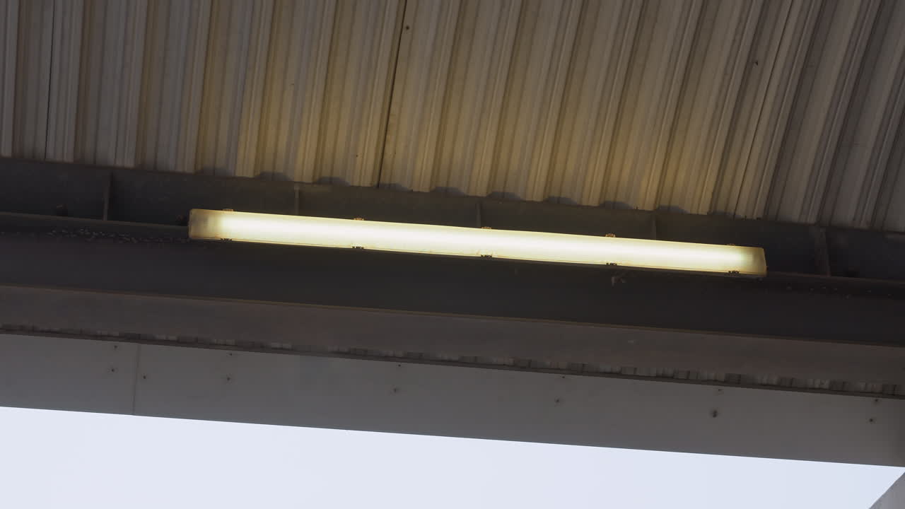 canopy fluorescent light under corrugated roof, steady warm glow across metal beam, subtle dirt and paint wear, daytime sky edge, functional service station atmosphere, maintenanceready mood, minimal