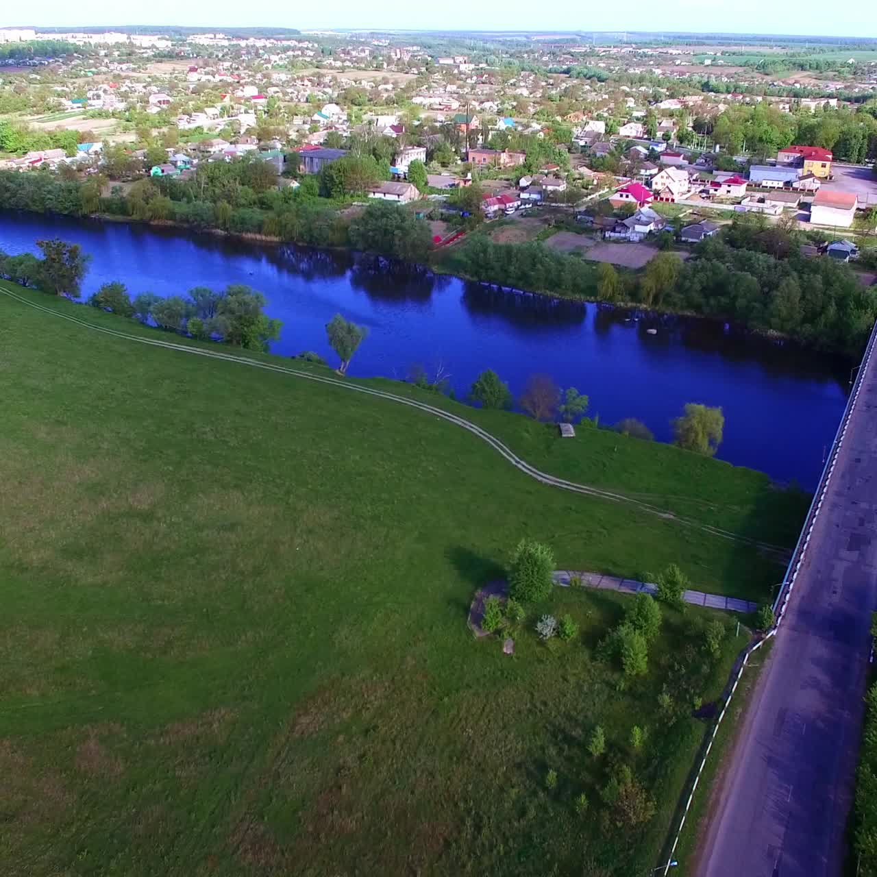 Blå elv som skiller landsbyen fra jordbruksland. Liten betongbro over elven. Solfylt bypanorama. Luftfoto