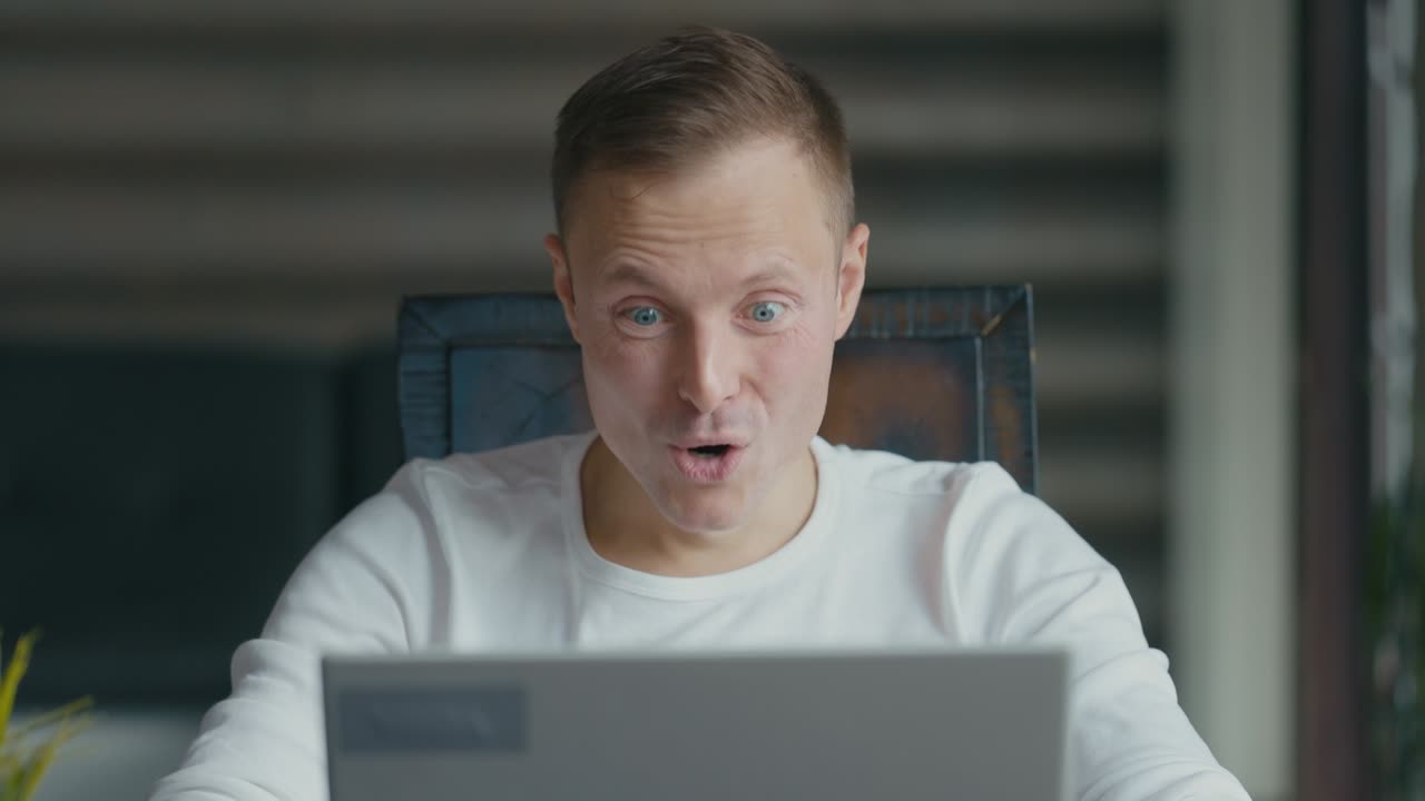 행복한 젊은이가 노트북 뒤에 있는 방에 앉아서 기쁜 소식을 듣고 기하며 '예'라고 말한다.