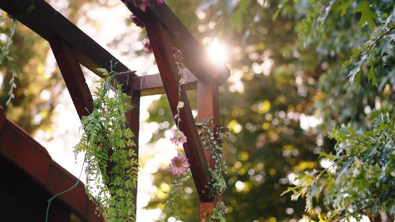 Wooden pergola hung with leafy garlands plus pink flowers for outdoor decoration, standing among tall trees on a sunny day