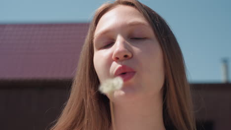 girl closes eyes as she gently blows dandelion puff under bright sunlight, holding delicate stem close to face with soft expression, framed by clear blue sky and rooftop in calm outdoor setting
