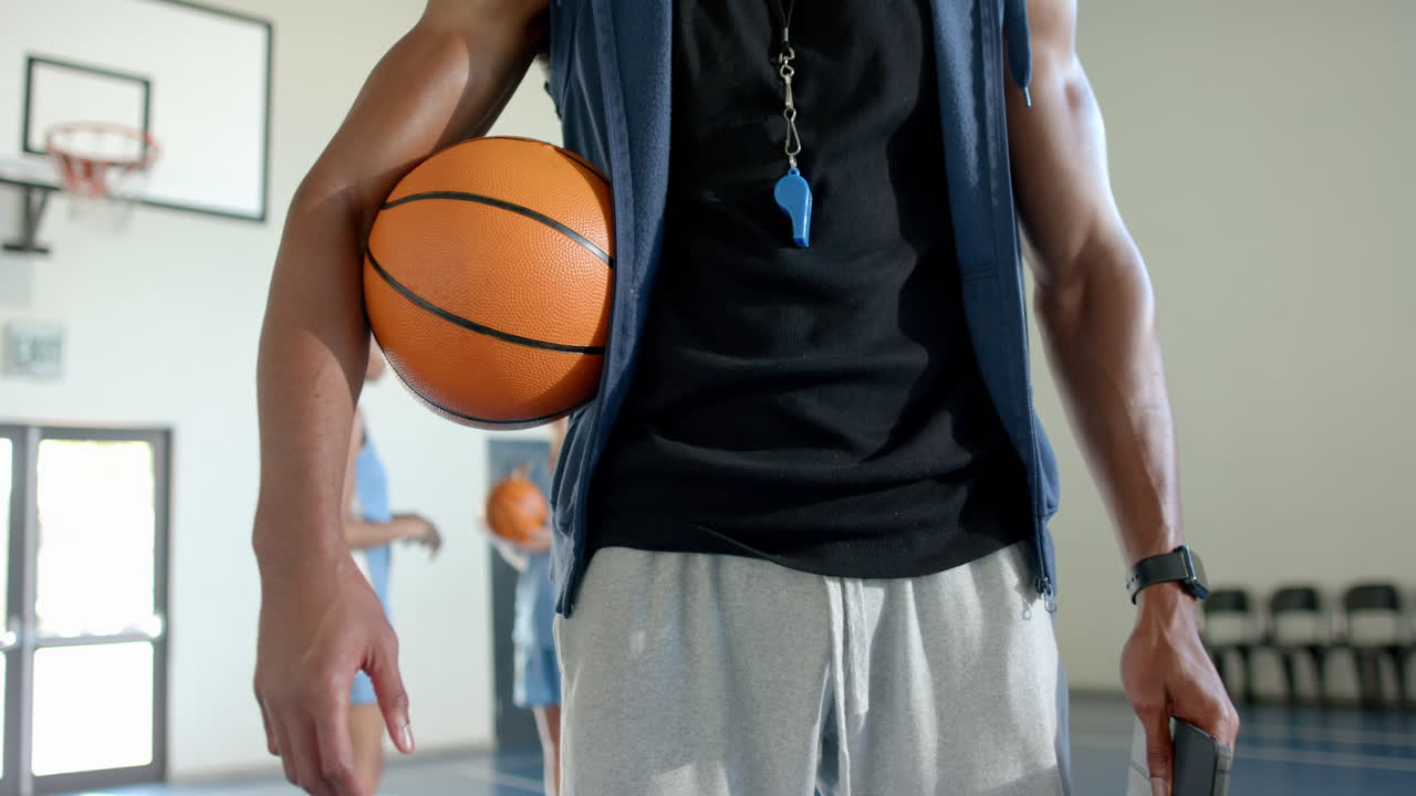 Holding basketball, coach with whistle standing in school gym, players practicing