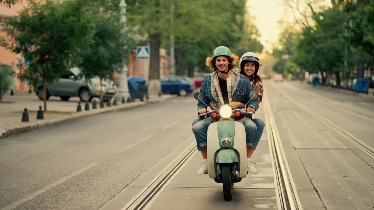 A happy couple, a guy with long curly hair in a denim jacket, rides with his happy girlfriend in a checkered shirt on a green moped with the headlight on along a wide summer street in the city