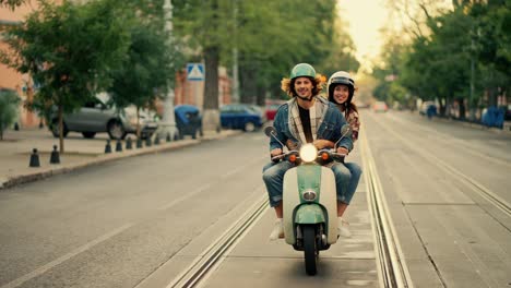 A-happy-couple,-a-guy-with-long-curly-hair-in-a-denim-jacket,-rides-with-his-happy-girlfriend-in-a-checkered-shirt-on-a-green-moped-with-the-headlight-on-along-a-wide-summer-street-in-the-city